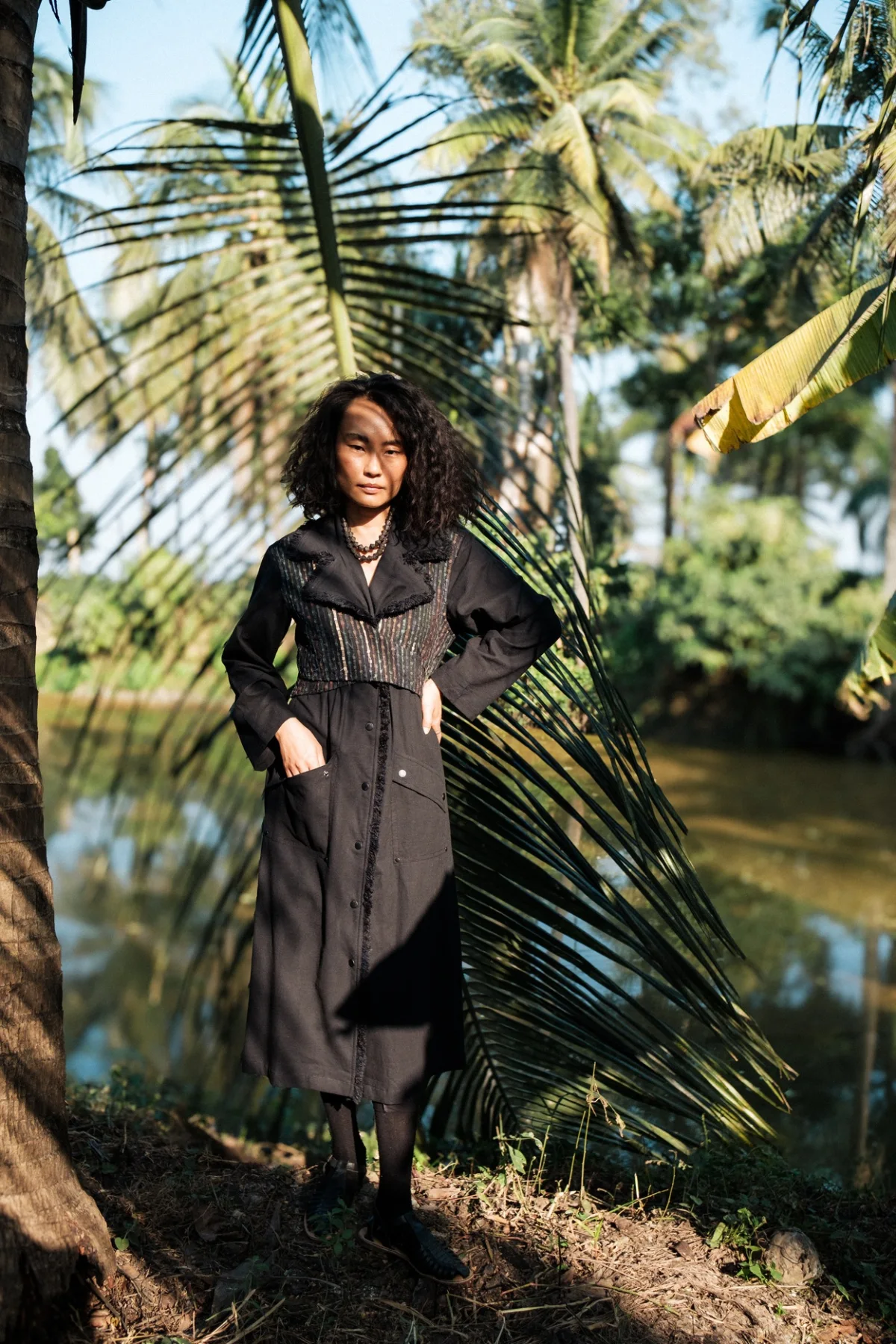 Model wearing a long dark coat beneath palm leaves